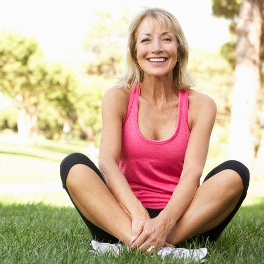 A smiling senior woman sitting cross-legged in grass wearing exercise clothes - The Body Firm